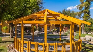 A wooden gazebo with a triangular roof, surrounded by a stone border, set in a landscaped garden with trees and a building in the background. - Serene Condo with pool, duck ponds and washer/dryer (Indio)