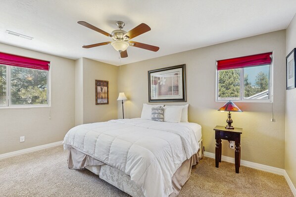 A bedroom with a bed, ceiling fan, two windows with red curtains, a bedside table with a lamp, and a framed picture on the wall.