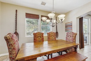 A dining room with a wooden table, four upholstered chairs, a chandelier, and a window with a view of trees.