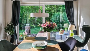 A dining area with a wooden table, green placemats, and a bowl. There are chairs, a vase with flowers, and a lamp. The room has large windows with curtains, and a view of greenery outside. - Cozy home in Lemvig with WiFi (Lemvig)