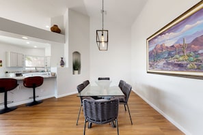 Una cocina moderna con armarios blancos y una isla central, un comedor con mesa de vidrio y sillas de mimbre, y un cuadro de paisaje grande en la pared.
