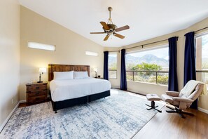 A hotel room with a large bed, a ceiling fan, a view of mountains through the window, a chair, and a nightstand with a lamp. - Expansive mountain views!  Spacious retreat near Catalina State Park! (Oro Valley)