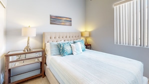 A bedroom with a large bed, a nightstand with a lamp, a wall-mounted painting, and a window with blinds. - Chic Cathedral City Home with Private Pool & Spa | Minutes to Palm Springs (Cathedral City)