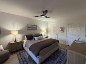 Een slaapkamer met een bed, twee nachtkastjes, een plafondventilator, een deur en een klein bureau met een lamp.