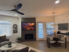 Ein modernes Wohnzimmer mit Flachbildfernseher, Deckenventilator, Kamin, Essbereich und einem großen Fenster mit Jalousien.