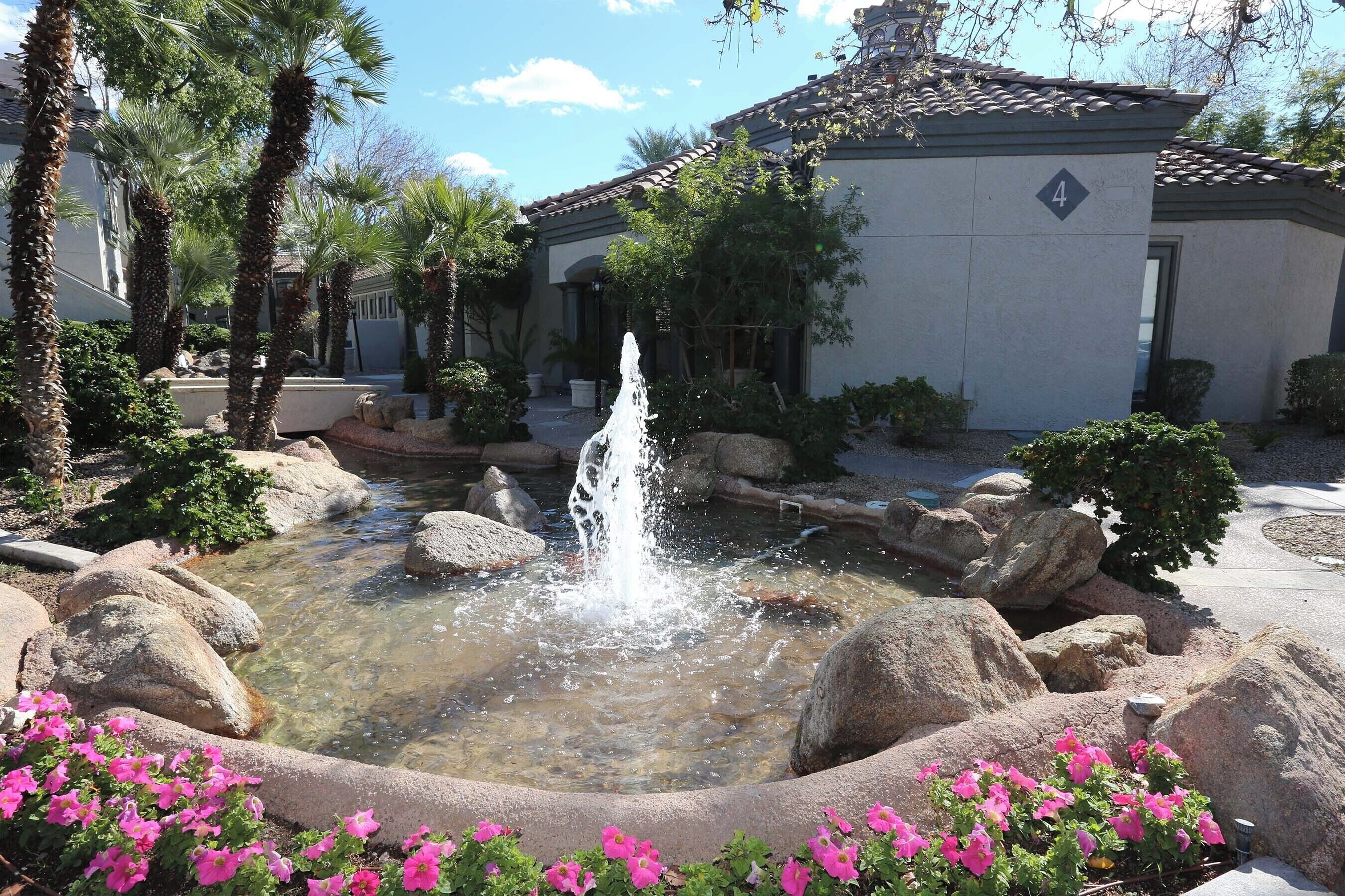 A courtyard with a water fountain, large rocks, and pink flowers.