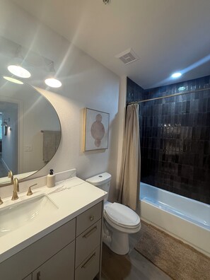 Een badkamer met een wit wastafelmeubel, een toilet en een douche met een donkere betegelde wand, plus een grote spiegel.