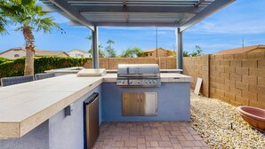 Et udendørs køkken med grill, vask og indbygget skab, omgivet af en terrasse med pergola.