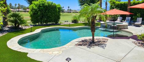 Piscina com borda de pedra, rodeada por um alpendre com cadeiras e guarda-sóis, e vista para um campo de golfe ao fundo.