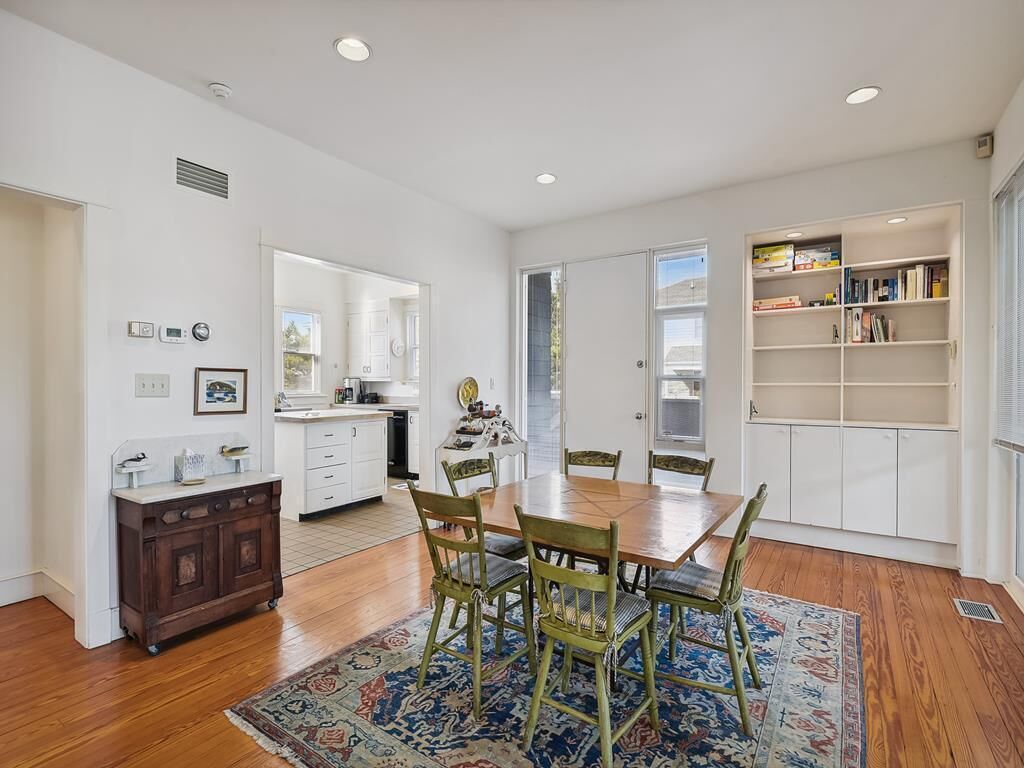 A spacious dining area with a wooden table and chairs, a patterned rug, and a white kitchen in the background.
