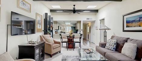 A spacious living room with a sofa, a coffee table, and a TV. There is a dining area with a table and chairs. The room has a ceiling fan and recessed lighting.