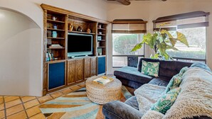 A living room with a television, wooden shelves, a sofa, and a large window with blinds.