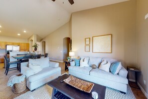 Ein geräumiger Wohnbereich mit grauem Sofa, einem Couchtisch und einem Essbereich, im Hintergrund ist eine Küche zu sehen.