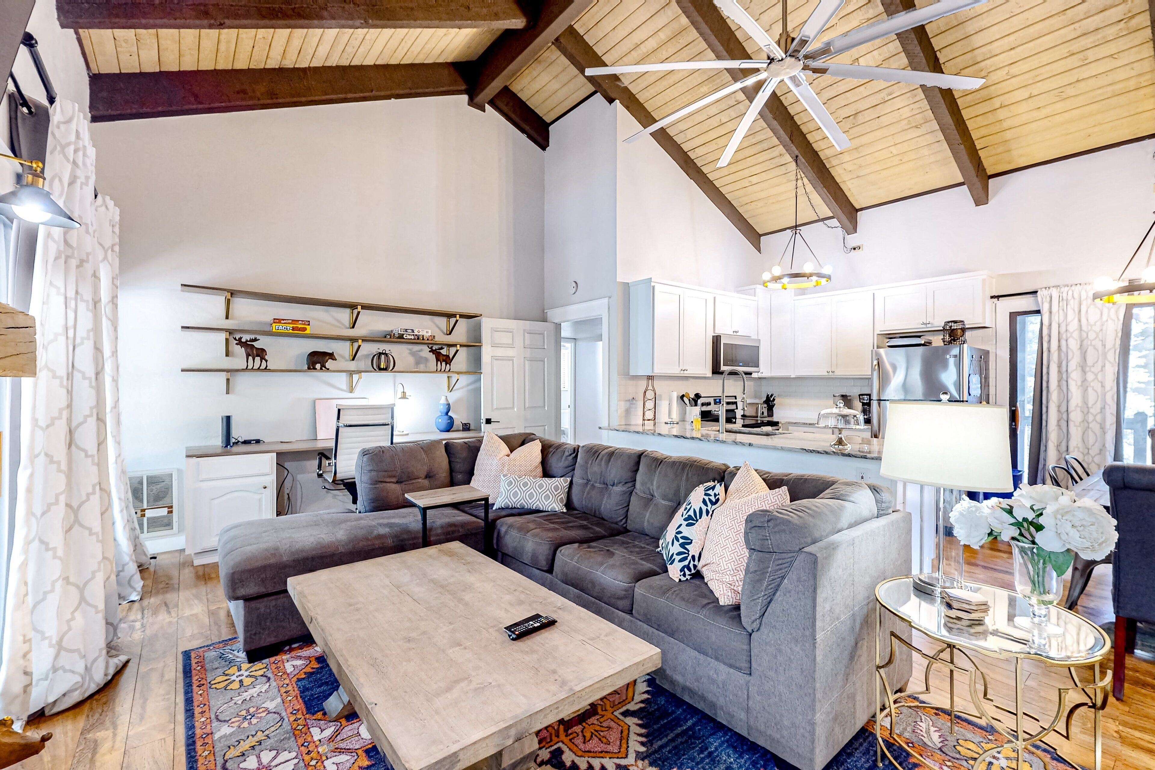 A spacious living room with a large gray sectional sofa, a wooden coffee table, and a ceiling fan. A kitchen with white cabinets and modern appliances is visible in the background.