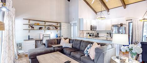 A spacious living room with a large gray sectional sofa, a wooden coffee table, and a ceiling fan. A kitchen with white cabinets and modern appliances is visible in the background.