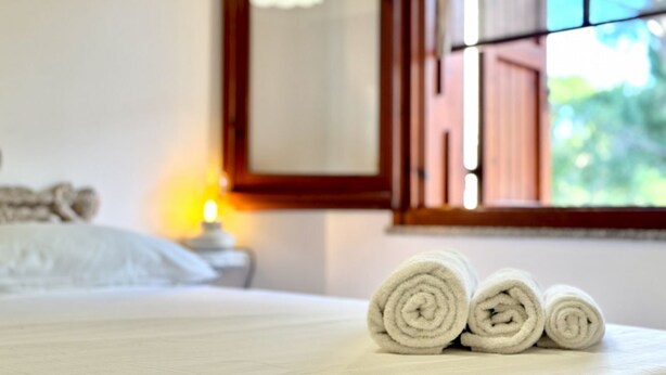 A neatly made bed with rolled white towels, a bedside lamp, and a window with a view of greenery. - Villa Prestige 🌊✨ - Refinement and Relaxation Just a Step from the Sea (Santa Margherita di Pula)