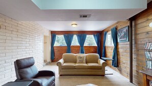 Ein gemütliches Wohnzimmer mit einer Ledersofa, einem beigefarbenen Sofa und einem kleinen Tisch. Große Fenster mit Vorhängen, eine Holzwand und ein Deckenlüfter sind ebenfalls vorhanden.