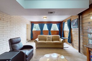 A cozy living room with a leather sofa, a beige couch, and a small table. There are large windows with curtains, a wooden wall, and a ceiling vent. - Shadow Mountain Village Escape | 2 Living Areas, Deck & Yard | Walk to Trail (Flagstaff)