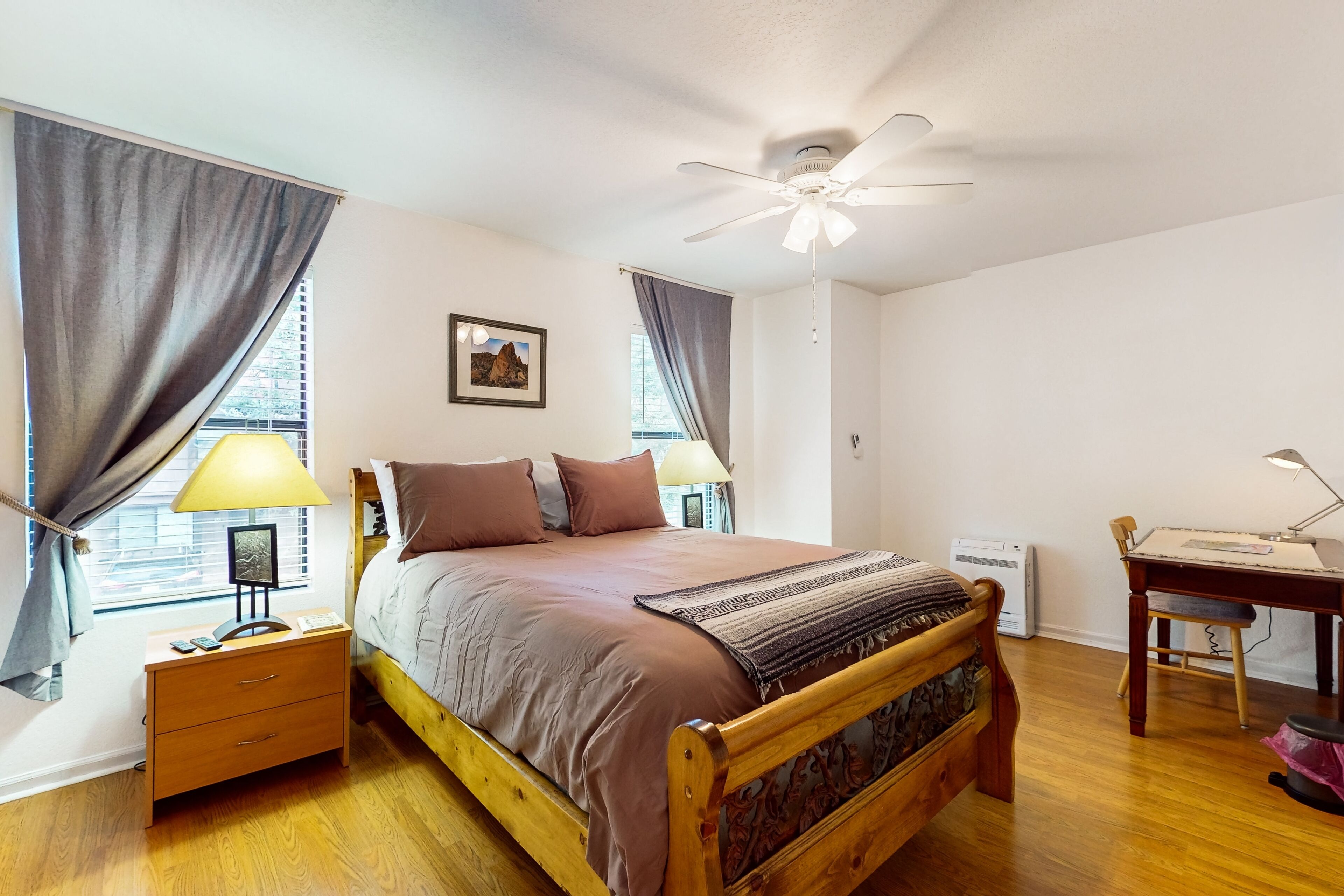 A bedroom with a wooden bed, a desk, a chair, a ceiling fan, and a window with curtains.