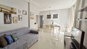 A modern living room with a grey sofa, a dining area with a white table and chairs, and a TV on a stand. - COAPI0100 - Edificio Palais Atlantique (Salvador)