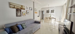 Ein modernes Wohnzimmer mit einem grauen Sofa, einem Essbereich mit einem weißen Tisch und Stühlen sowie einem Fernseher auf einem Schrank.