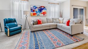 A living room with a sectional sofa, a blue armchair, a patterned rug, and a large abstract painting on the wall. - Charming Condo with Central A/C & Washer/Dryer - Shared Pool & Hot Tub (Phoenix)