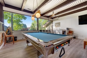 A pool table with a built-in ball rack, a ceiling fan, and a window with a view of trees. - Refined home with a game room, balcony, grill, washer/dryer, & four bedrooms (Flagstaff)