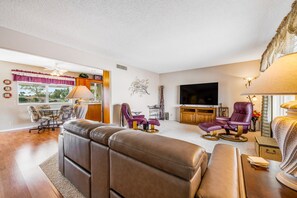 Ein Wohnzimmer mit einer Eckcouch, zwei violetten Sesseln, einem Holztisch, einem Fernseher und einem Essbereich mit Tisch und Stühlen.