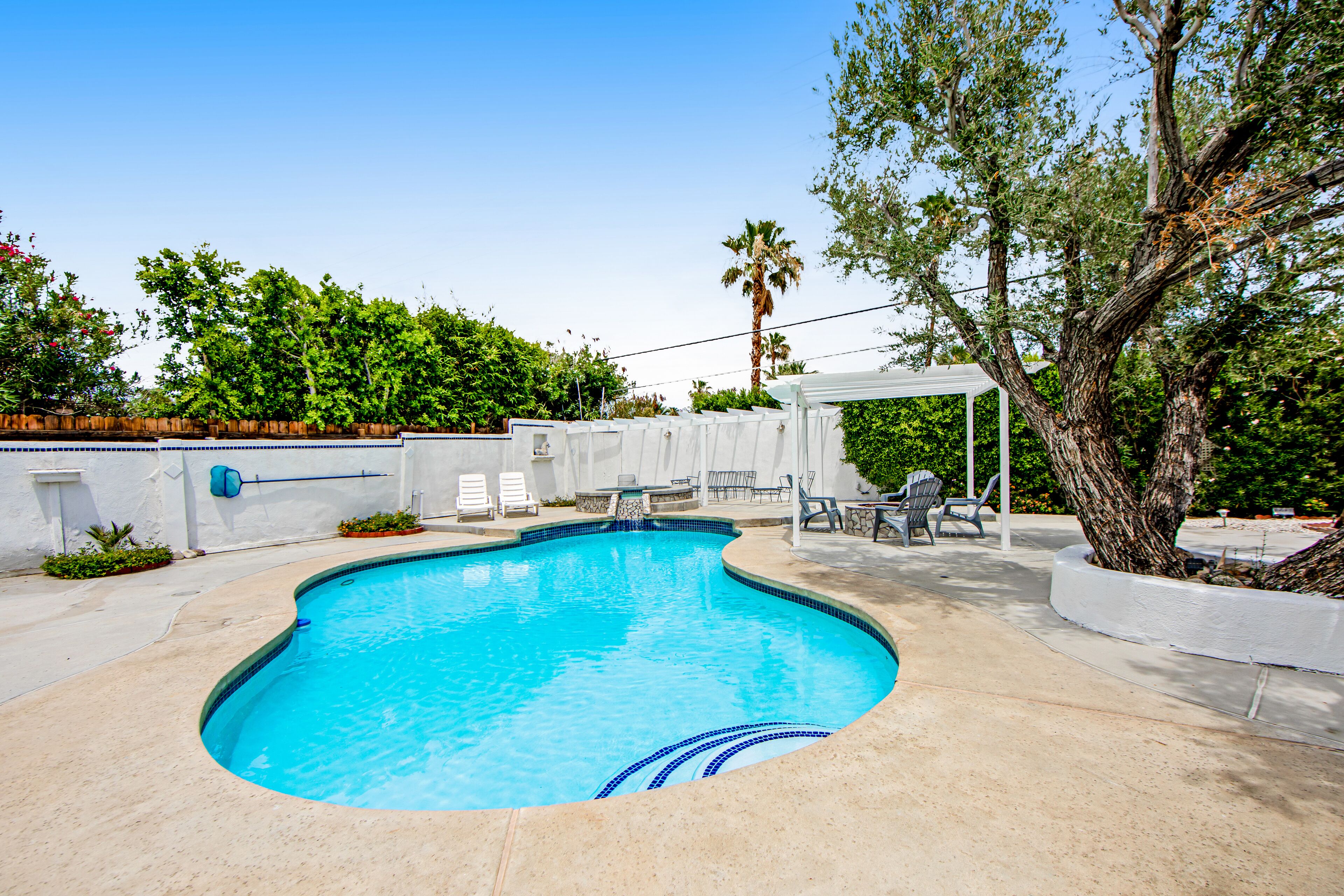 Une piscine à bordures courbes, entourée d’une terrasse en béton. On y trouve des chaises longues et un espace patio couvert avec une table et des chaises.