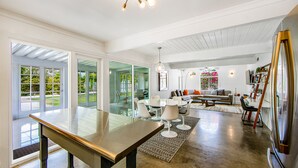 A spacious living area with a glass dining table, modern seating, and a large window offering a view of greenery. - Appealing Racquet Club home with firepit, private pool & gas grill (Palm Springs)