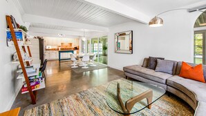A spacious living area with a glass coffee table, a sofa with cushions, a bookshelf, and a kitchen in the background. - Appealing Racquet Club home with firepit, private pool & gas grill (Palm Springs)