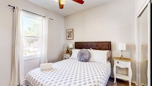 Un dormitorio con cama, ventilador de techo, ventana con cortinas, mesita de noche con lámpara y un cuadro enmarcado en la pared.