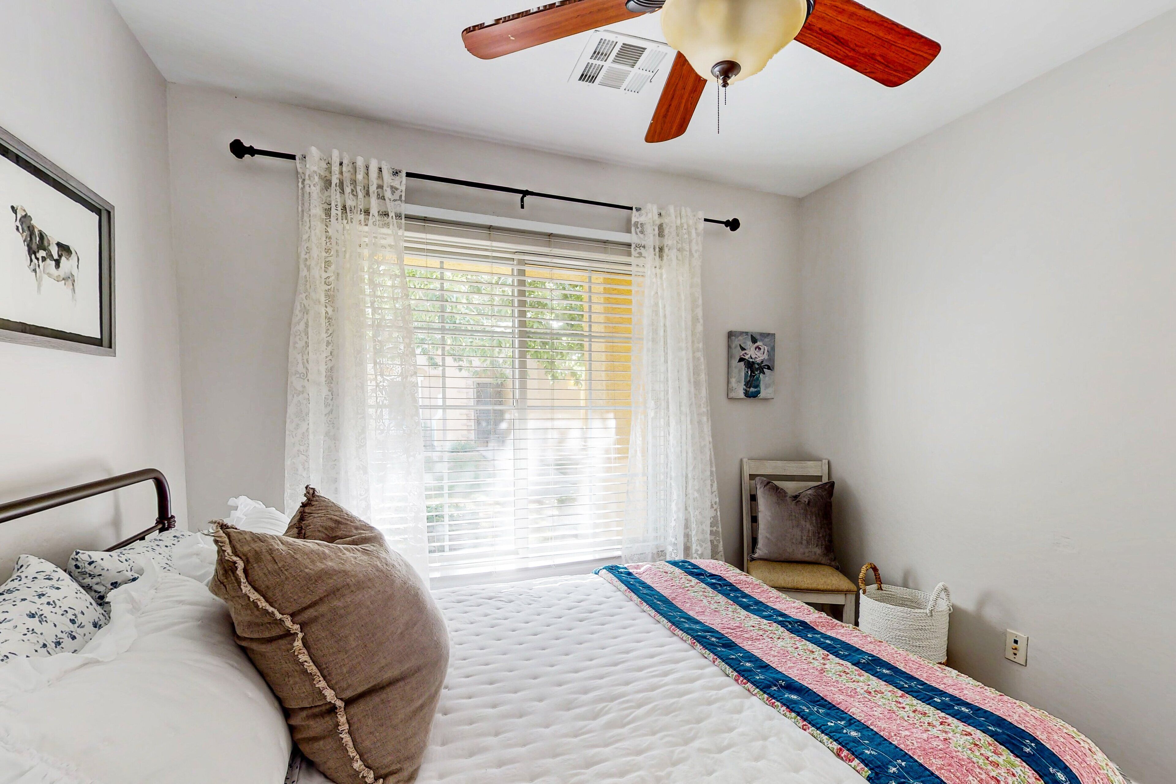 Un dormitorio con cama, ventilador de techo, ventana con cortinas, mesita de noche y estante en la pared.
