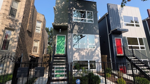 Un edificio moderno con una escalera que lleva a una puerta verde, rodeado por una cerca de metal negro y una acera con flechas amarillas indicando dirección.