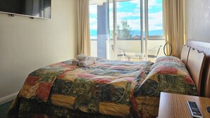 Habitación de hotel con una cama grande, un televisor montado en la pared, un balcón con vistas al cielo y edificios, y una pequeña mesa con un control remoto.
