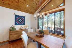 A dining area with a wooden table, white chairs, a chandelier, and a large window with a view of trees. - Luxurious mountain home w/ firepit & grill - close to hiking, fishing, & golf (Show Low)