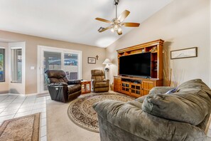 A living room with a large flat-screen TV, leather recliners, a ceiling fan, and a wooden entertainment center. - Lovely getaway with private pool, putting green & desk (Gilbert)