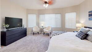 A bedroom with a bed, a TV, a dresser, two chairs, a small table, and three windows with blinds.