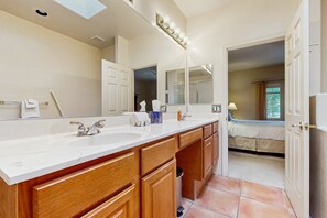 A bathroom with a large vanity, two sinks, a mirror with lights, and a view into a bedroom.