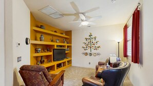 Un salon avec une bibliothèque jaune, un téléviseur, un ventilateur de plafond et un élément décoratif mural.