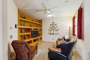 A living room with a yellow bookshelf, a television, a ceiling fan, and a decorative wall piece. - Comfortable getaway with hot tub, pool, private BBQ & full kitchen (Oro Valley)