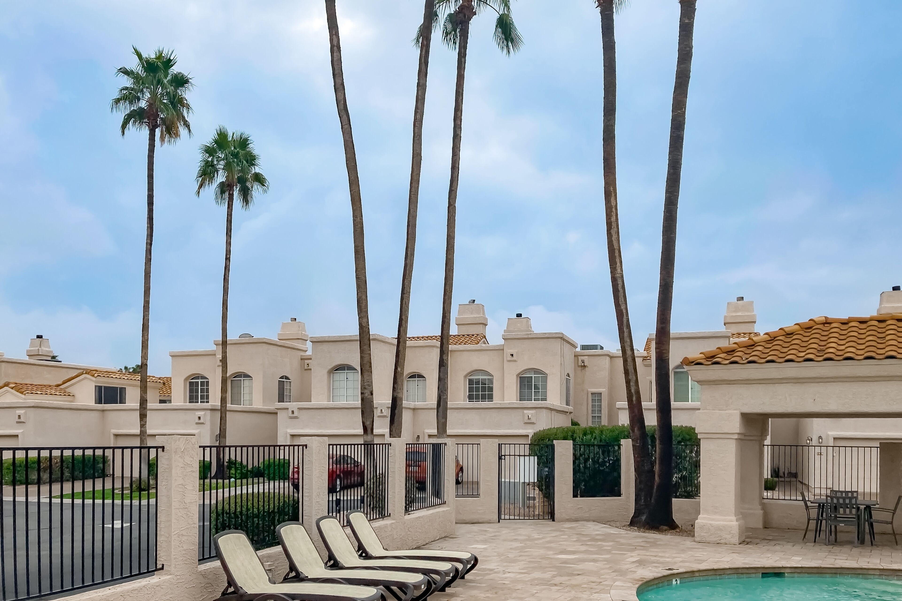 Zona de piscina con sillones de descanso, palmeras y un edificio con tejado de tejas.