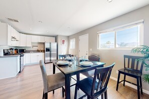 A modern kitchen with white cabinets, a dining table set for four, and a large window providing a view of the outdoors. - Well-equipped modern home with mountain views, washer/dryer, full kitchen, & AC (Joshua Tree)