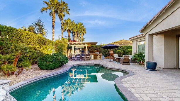 Ein Pool mit gewölbter Kante, umgeben von Palmen und einer Terrasse mit Gartenmöbeln.