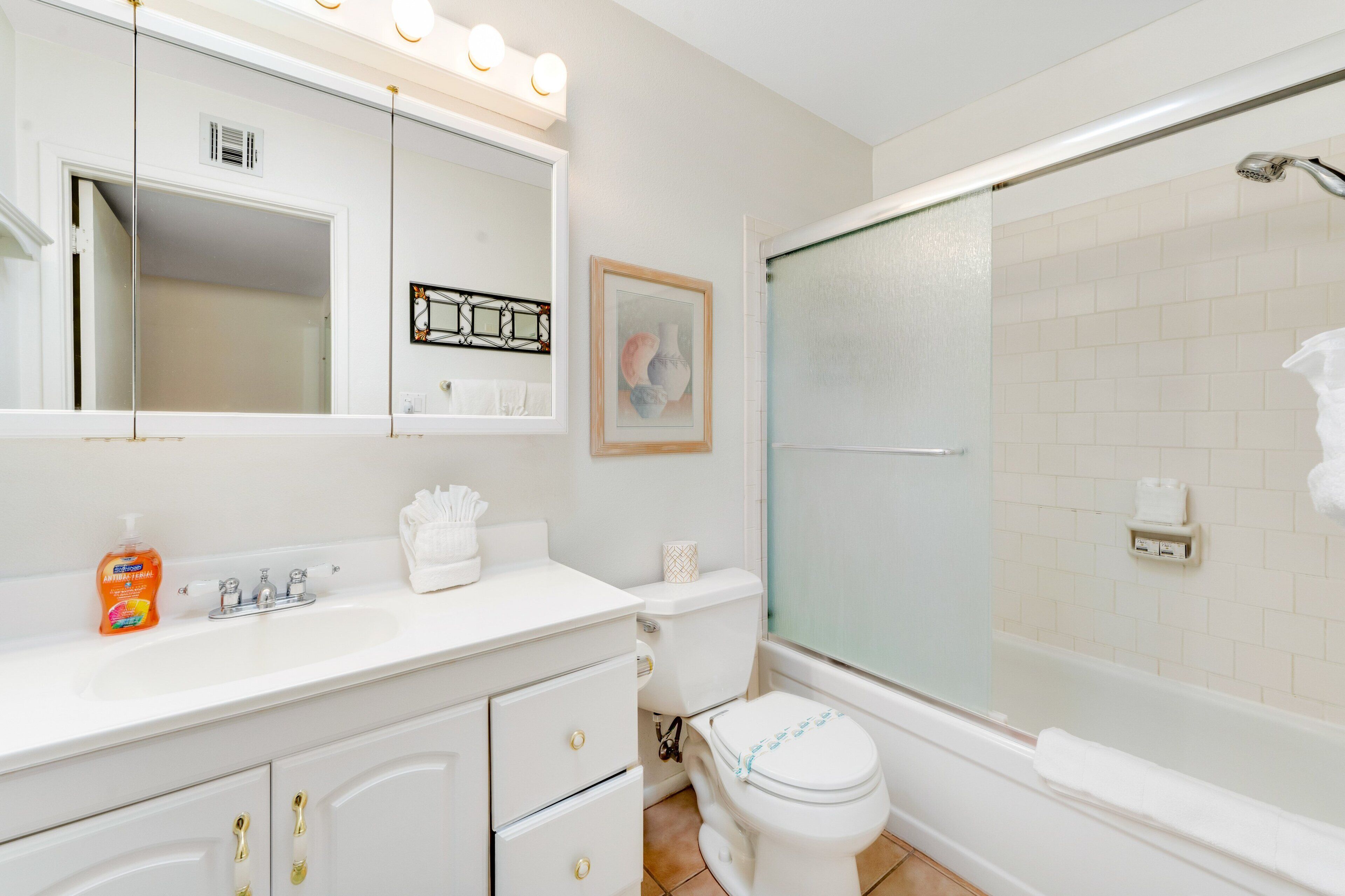 A bathroom with a white sink, mirror, and toilet, a shower with a glass door, and a framed picture on the wall.