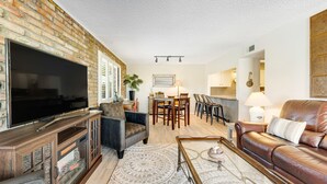 Un salon avec un mur en briques, un téléviseur à écran plat, un canapé en cuir, un coin repas avec une table et des chaises, et une cheminée.