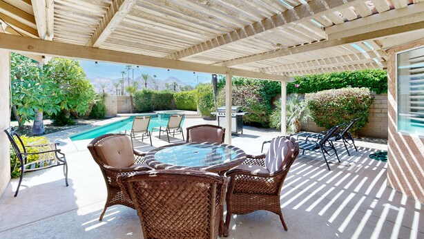 A covered patio area with wicker furniture and a glass-top table, overlooking a swimming pool and greenery. - Beautiful home with private pool, enclosed backyard & mountain view (Indian Wells)