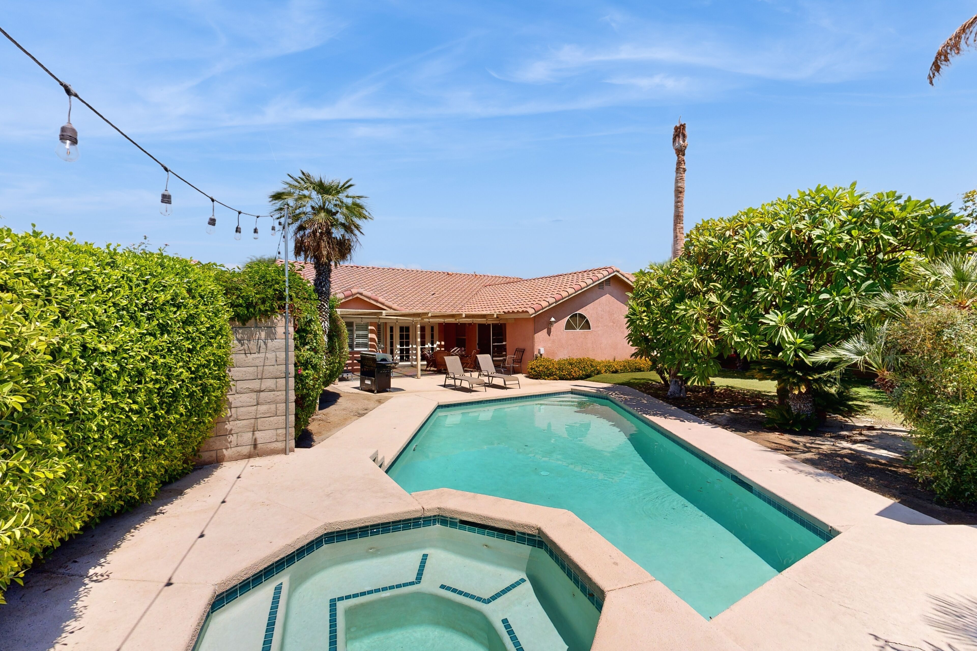 A swimming pool with a hot tub, surrounded by greenery and a house with a red-tiled roof.