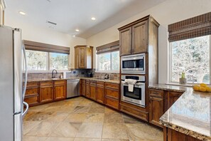A spacious kitchen with wooden cabinets, a stainless steel refrigerator, a built-in oven, and a large window with a view of trees.