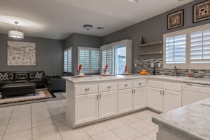 A modern kitchen with white cabinets, a large island, and a dining area with a black sofa. - Mountain view home with pool, spa and dog friendly (La Quinta)